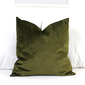 May include: A square throw cushion covered in a solid olive green velvet fabric. The cushion features a soft, plush texture with a slight sheen and a small black fabric tag visible on the lower left side, presenting a clean and uniform appearance.