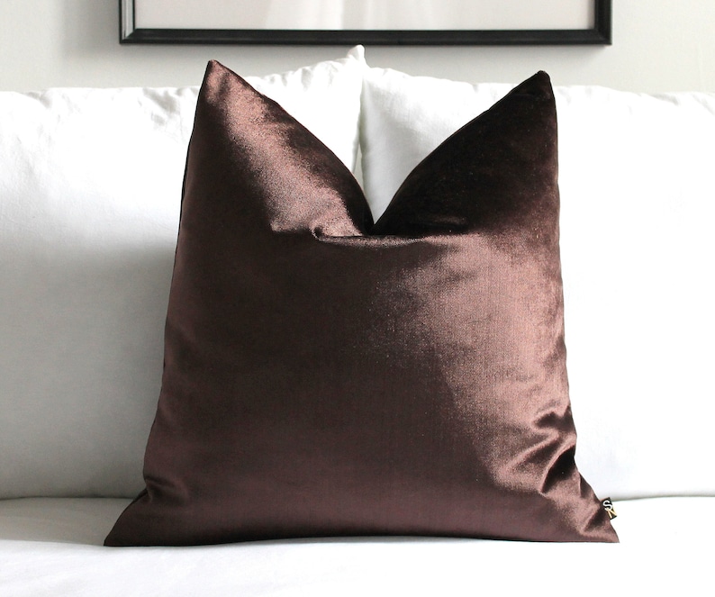 K&ouml;nnte beinhalten: Ein quadratisches Kissen aus schokoladenbraunem Samt. Das Kissen hat einen weichen Glanz und liegt auf einer wei&szlig;en Oberfl&auml;che. Das Kissen ist ein dekoratives Element f&uuml;r ein Bett oder Sofa.