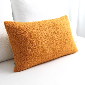 May include: Mustard-colored rectangular pillow with a textured, plush surface. The pillow is placed on a white surface, with a white pillow visible in the background. The pillow's color is a warm, inviting shade.