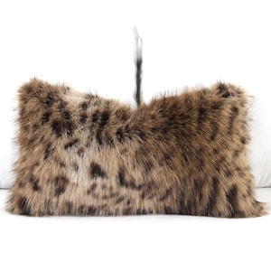 May include: A rectangular decorative pillow with a faux fur cover. The pillow features a brown and tan spotted pattern, resembling animal fur. The pillow is placed on a white surface.
