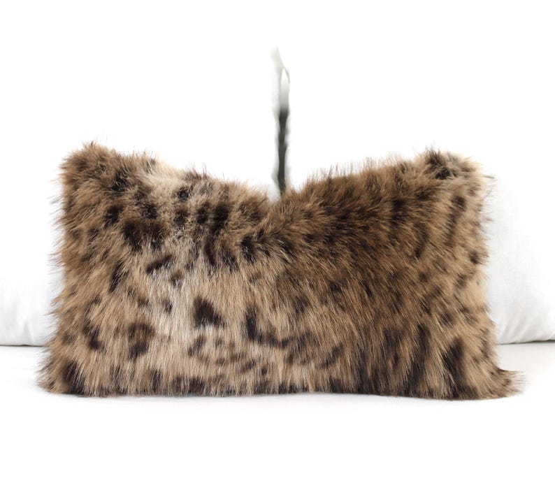 May include: A rectangular decorative pillow with a faux fur cover. The pillow features a brown and tan spotted pattern, resembling animal fur. The pillow is placed on a white surface.