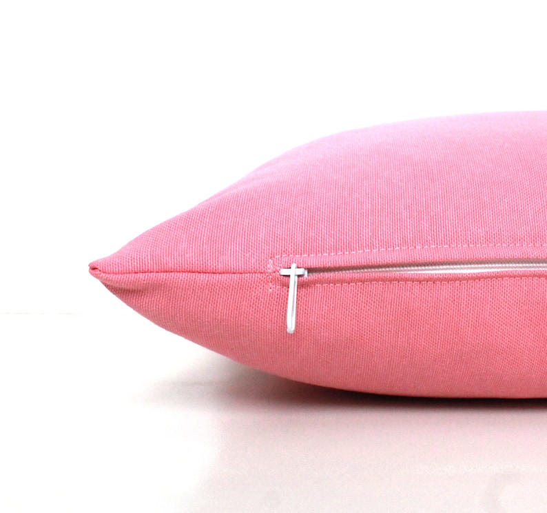 Puede incluir: Una almohada rosa con una cremallera blanca. La almohada est&aacute; hecha de una tela suave y texturizada.