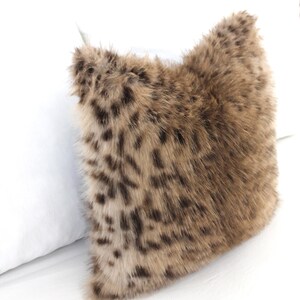 May include: A square decorative pillow with a faux fur cover. The pillow features a brown and tan spotted pattern, resembling a leopard print. The pillow is placed next to a white pillow.