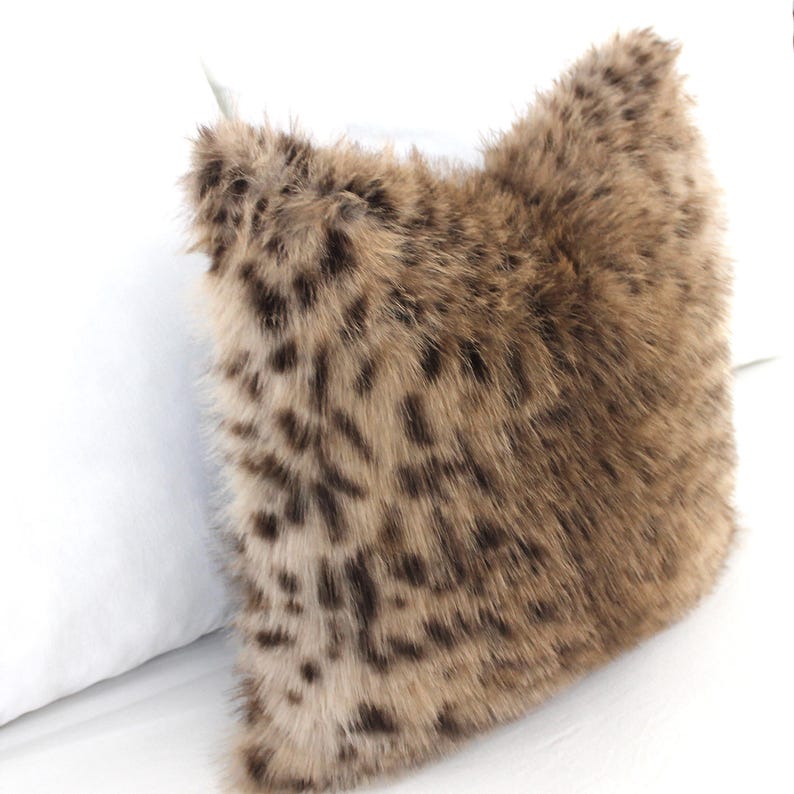 May include: A square decorative pillow with a faux fur cover. The pillow features a brown and tan spotted pattern, resembling a leopard print. The pillow is placed next to a white pillow.