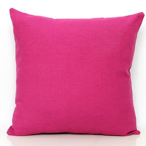 May include: A square magenta cushion with a solid colour design. The cushion has a soft, textured appearance and is set against a white background. The cushion is a vibrant shade of pink.