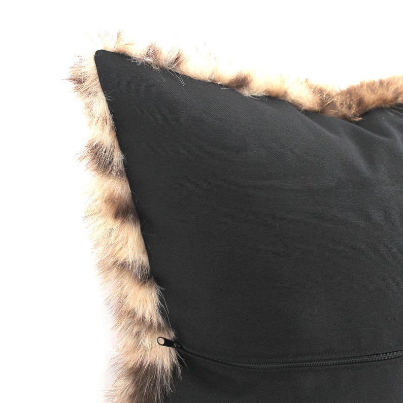 May include: A black pillow with a faux fur trim in shades of brown and tan. The pillow has a zipper closure along one edge. The pillow is a decorative item for a bed or sofa.