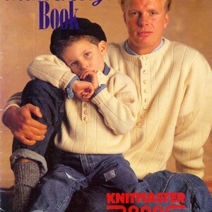 May include: A father and son wearing cream colored sweaters with button details. The father is wearing blue jeans and the son is wearing blue jeans with a black beret. The text "The Mens & Boys Book" is at the top of the image. The text "Knitmaster 7000 creative kone 4ply Aneto Blackjack" is at the bottom of the image.