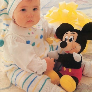 Pode incluir: Um bebê usando um chapéu de tricô branco e verde listrado e um macacão de tricô branco com bolinhas coloridas. O bebê está segurando um brinquedo de pelúcia do Mickey Mouse. O texto "Lots for Tots in 3 ply Feathersoft" está na parte inferior da imagem.