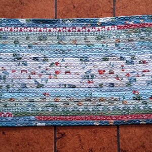 Può includere: Tappeto ovale intrecciato con un mix di tessuti blu, verdi e rossi. Il tappeto presenta una sezione centrale con sfondo bianco e un motivo di piccoli fiori rossi e blu. I bordi sono bordati con vari tessuti fantasia.