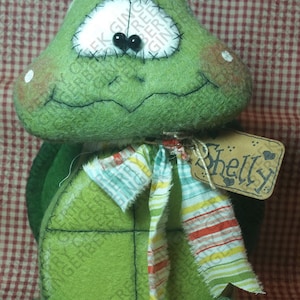 Może przedstawiać: Ręcznie wykonana zielona pluszowa zabawka w kształcie żółwia z szytą twarzą, czarnymi koralikowymi oczami i kolorową, pasiastą szarfą. Żółw ma metkę z napisem "Shelly". Pluszak znajduje się na czerwono-białym kratkowanym tle.