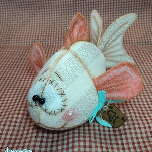 Pode incluir: Um brinquedo de peixe de feltro feito à mão com corpo creme, barbatanas pêssego e um rosto costurado. Apresenta olhos de contas pretas e uma pequena etiqueta amarrada com uma fita azul. A etiqueta diz "Git".