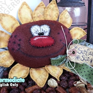 May include: A handmade, quirky sunflower decoration with a smiling face, red nose, and white eyes. The sunflower has tan petals and a tag that reads "Sunflower Sue". The decoration is made of felt and fabric.