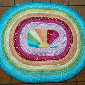 May include: Oval braided rug with a rainbow of colors, including blue, yellow, orange, and pink. The center features a sunburst design in various patterned fabrics. A decorative floor covering.