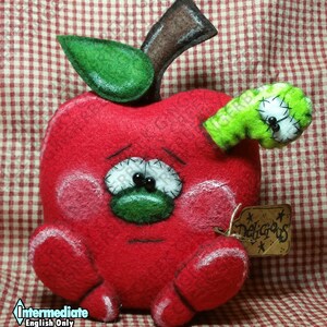 May include: A plush red apple with a sad face, green stem and leaf, and a green worm. The apple has white stitched eyes, pink cheeks, and a tag that says "Delicious". The apple is on a red and white checkered background.