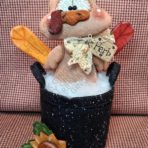 May include: A handcrafted Thanksgiving turkey decoration. The turkey is made of felt and sits in a black pot with a sunflower accent. The turkey has a tag that says "Herb". The background is a red and white checkered pattern.