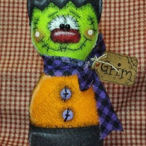 May include: A handmade Frankenstein's monster doll with a green face, black hair, and a purple plaid scarf. The doll has an orange body, black eyes, and a red mouth. It has a tag that says "Grim".