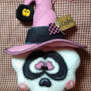 May include: A handmade Halloween decoration featuring a skull wearing a pink witch hat with a black brim. The skull has black eyes and stitched details. A tag reads "D. Ceased". The decoration is on a red and white checkered background.