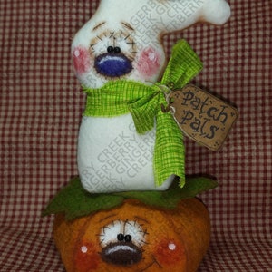 Puede incluir: Decoración de Halloween hecha a mano con una figura de fantasma blanco sobre una calabaza naranja. El fantasma tiene una bufanda verde y una etiqueta que dice "Patch Pals". La calabaza y el fantasma tienen rasgos faciales cosidos y mejillas rosadas.