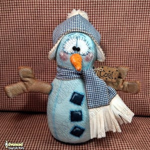 Könnte beinhalten: Handgefertigte Schneemannpuppe mit hellblauem Körper, mit blau-weiß kariertem Hut und Schal. Sie hat eine Karottennase, schwarze Knopfaugen und braune Stockarme. Ein Etikett lautet "Coal".