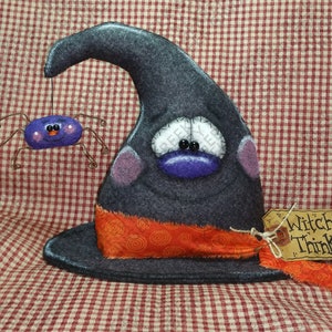 Könnte beinhalten: Eine skurrile Halloween-Dekoration mit einem grauen Hexenhut mit lila Gesicht und orangefarbenem Band. Eine lila Spinne hängt am Hut. Das Etikett lautet "Witchful Thinking".