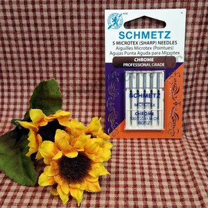 Puede incluir: Un paquete de agujas SCHMETZ Microtex afiladas, con texto en ingl&eacute;s, franc&eacute;s y espa&ntilde;ol. El paquete es naranja, morado y blanco. La imagen tambi&eacute;n incluye un grupo de girasoles amarillos con hojas verdes.