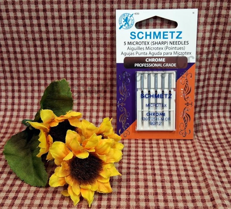 Puede incluir: Un paquete de agujas SCHMETZ Microtex afiladas, con texto en ingl&eacute;s, franc&eacute;s y espa&ntilde;ol. El paquete es naranja, morado y blanco. La imagen tambi&eacute;n incluye un grupo de girasoles amarillos con hojas verdes.