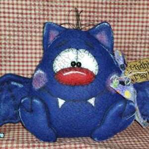 Op de afbeelding: Een donkerblauwe vilten vleermuis met grote witte ogen, een rode mond en witte hoektanden. De vleermuis heeft grote vleugels en een label met de tekst "Midnite Mayhem". De achtergrond is een rood-wit geblokt patroon.