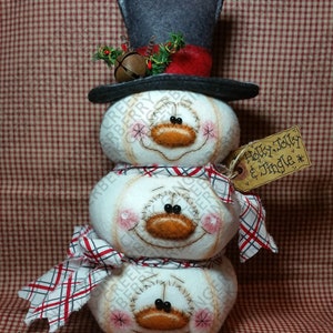 Op de afbeelding: Een handgemaakte sneeuwmanversiering met een hoge hoed, sjaal en een label met de tekst "Holly, Jolly & Jingle". De sneeuwman is gemaakt van drie gestapelde witte bollen met gestikte gezichtskenmerken, roze wangen en een oranje wortelneus. De sjaal heeft een rood-wit ruitpatroon.