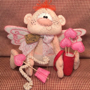 Peut inclure: Poupée Cupidon faite à la main, corps rose, ailes et nœud papillon. Elle tient un arc et une flèche, et une carquois de flèches en forme de cœur. La poupée porte une étiquette «Cupidon». Sur fond à carreaux rouges et blancs.