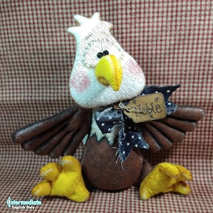 Może przedstawiać: Ręcznie wykonana patriotyczna figurka orła z białą głową, żółtym dziobem i brązowymi skrzydłami. Orzeł ma niebiesko-białą muchę w gwiazdki i etykietę z napisem "Noble". Stopy orła są żółte, na tle czerwono-białej kraty.