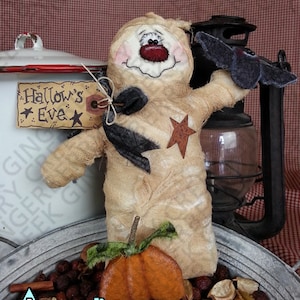 May include: A handcrafted Halloween doll with a painted face, holding a felt bat. The doll has a burlap-style body with a star and black accents. A small pumpkin and a sign that says "Hallow's Eve" are also present.