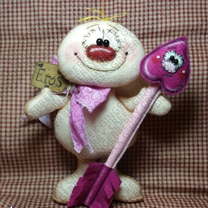 May include: A handmade felt doll with a smiling face, holding a pink heart-shaped arrow. The doll has a pink scarf and a tag that says "Eros". The background is a red and white checkered pattern. The doll's height is approximately 8 inches.