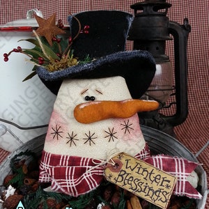 Könnte beinhalten: Handgefertigte Schneemann-Dekoration mit schwarzem Zylinder, orangefarbener Karottennase und rot-weiß kariertem Schal. Der Schneemann hat gestickte Schneeflocken und das Etikett lautet "Winter Blessings". Der Schneemann befindet sich in einem Metallbehälter mit Grünzeug.