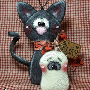 Może przedstawiać: Ręcznie robiona dekoracja na Halloween z filcu przedstawiająca kota i ducha. Szary kot ma różowy nos i czerwoną i pomarańczową wstążkę. Duch jest biały z szarymi oczami i różowymi policzkami. Etykieta głosi "Risky".