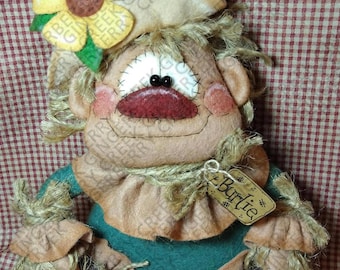May include: A handmade scarecrow doll with a tan face, stitched features, and a large red mouth. It has a yellow sunflower on its head and wears a green shirt. The doll has a tag that says "Burlie".