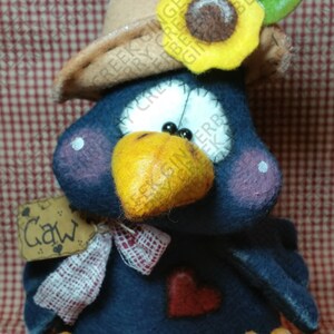 May include: A handcrafted, plush bird figure in navy blue, wearing a tan hat adorned with a yellow sunflower and green leaves. It has a yellow beak and feet, a red heart, and a tag that says "Gaw".