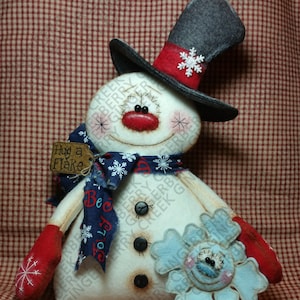 May include: A handmade snowman doll with a top hat, scarf, and mittens. The snowman has a red nose, stitched eyes, and a snowflake on its hat. It holds a blue snowflake and a tag that says "Hug a Flake".