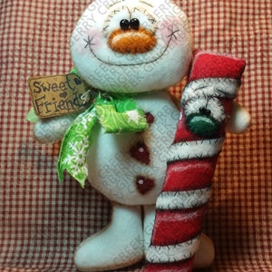 May include: A handmade snowman doll with a carrot nose, black button eyes, and a stitched smile. It holds a red and white candy cane and wears a green scarf. A tag reads "Sweet Friends."