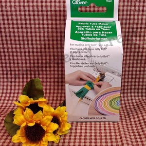 Puede incluir: Una caja de producto Clover Fabric Tube Maker, con texto en varios idiomas. La caja es blanca y verde, con una foto de la herramienta en uso. Un grupo de girasoles amarillos se encuentra frente a la caja sobre una superficie a cuadros rojos y blancos.