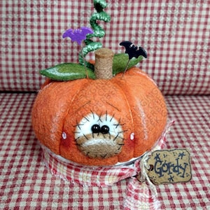 May include: A felted orange pumpkin figurine with a tan muzzle, black bead eyes, and stitched eyelashes. It features a green felt leaf, a brown stem, a green curly vine, and two bat-shaped pins. A red and white plaid fabric strip wraps the base.
