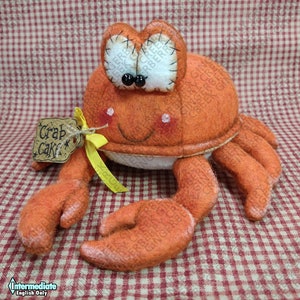 May include: An orange felt crab plush toy with large, stitched eyes and a smiling face. It has a tag that reads "Crab Cake" tied with a yellow ribbon. The crab is on a red and white checkered background.