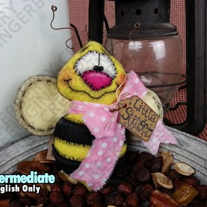 May include: A handmade bee-shaped ornament with a yellow head, black and yellow striped body, and pink bow. The ornament has a stitched face and a tag that reads "Little Stinger". The bee sits in a metal container with dried fruit.
