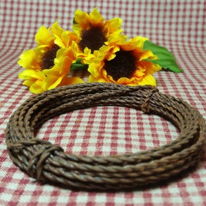 May include: A coiled, brown rope bracelet with a rustic appearance. The bracelet is made of tightly wound strands, creating a textured surface. Bright yellow sunflowers with brown centers and green leaves are in the background, set against a red and white checkered pattern.