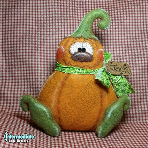 Könnte beinhalten: Eine handgemachte orangefarbene Kürbispuppe mit grünem Stiel und Füßen, die ein grünes Bandana trägt. Die Puppe hat aufgenähte Gesichtszüge und ein Etikett mit der Aufschrift "Jack". Die Puppe sitzt auf einer rot-weiß karierten Oberfläche.