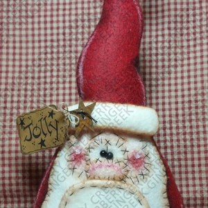 May include: A festive Christmas ornament depicting Santa Claus. It features a red hat with a gold bell, a white beard, and a tag reading "Jolly". The face is detailed with stitched features and pink cheeks.