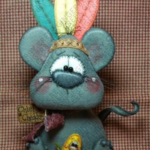 May include: A handmade gray mouse doll with a Native American headdress made of colorful feathers. The doll has a small sign that says "Chief Cheddar" and is holding a corn on the cob. The background is a red and white checkered pattern.