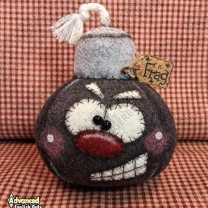 May include: A handmade, dark gray felt bomb with a cartoon face, white stitched eyes, and a red mouth. It has a gray cap, a white tassel, and a small tag that says "Frag". The bomb is sitting on a red and white checkered surface.