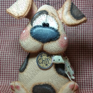 Può includere: Un cane di peluche fatto a mano con dettagli beige, marroni e neri. Il cane ha un naso blu, occhi neri e un'etichetta con la scritta "Chip". Le orecchie e le zampe hanno macchie più scure. Il cane è su una superficie a scacchi rossi e bianchi.
