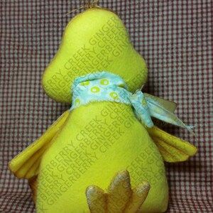 May include: A handmade yellow duck plush toy with a light blue and white patterned scarf. The duck has a large head, small wings, and a detailed foot. The background is a red and white checkered pattern.