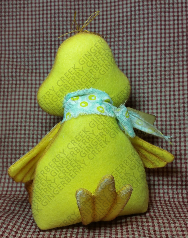 May include: A handmade yellow duck plush toy with a light blue and white patterned scarf. The duck has a large head, small wings, and a detailed foot. The background is a red and white checkered pattern.